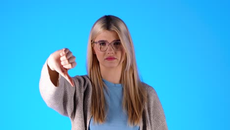 Disappointed-young-woman-shows-thumbs-down,-isolated-blue-background,-studio