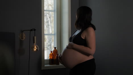 Pregnant-woman-in-third-trimester-standing-by-window,-hand-on-belly,-intimate-mood