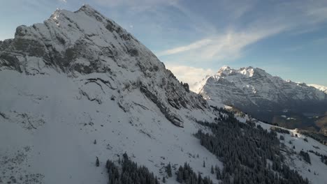 Backlit-swiss-alps-mountain-with-pine-tree-forest-on-slope-of-sharp-steep-cliffs,-epic-light-on-wispy-cloud