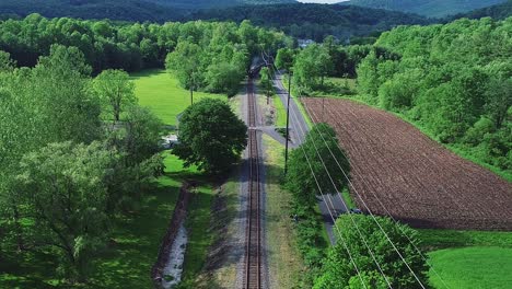 Una-Vista-Aérea-De-Un-Largo-Tren-De-Pasajeros-De-Vapor-Acercándose-En-Una-Sola-Vía-Viajando-A-Través-De-Verdes-Tierras-De-Cultivo-En-Un-Día-De-Primavera