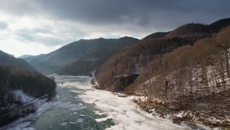 Siriu-lake-in-winter,-with-surrounding-bare-trees-and-snowy-banks,-aerial-view