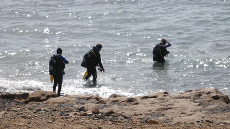 Buzos-Con-Elegantes-Equipos-De-Buceo-Se-Embarcan-Desde-Una-Playa-Rocosa,-Desafiando-Las-Brillantes-Olas-En-Una-Emocionante-Aventura-En-El-Océano.