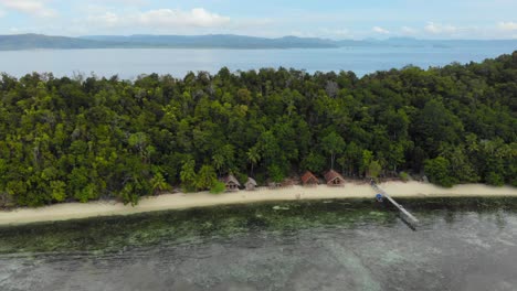 Perfectly-clear-water-showing-turquoise-ocean-floor-in-front-of-island-paradise-of-Kri-in-Raja-Ampat,-Indonesia-with-palm-trees-and-vacation-huts,-aerial-dolly-out