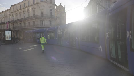 Straßenreiniger-Zu-Fuß-In-Montpellier,-Blaue-Straßenbahn-Hielt-An,-Im-Hintergrund-Ein-Wunderschöner-Sonnenuntergang-Und-Historische-Gebäude