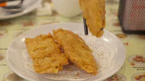 Chopsticks-picking-up-fried-tofu-skin-spring-roll-from-plate,-close-up