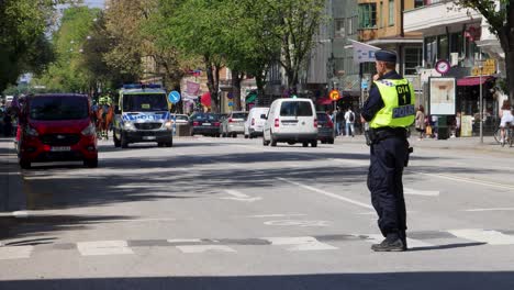 Police-officer-directing-traffic-on-Sveavägen,-Stockholm,-sunny-day,-with-emergency-vehicles-in-the-background
