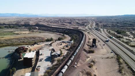 Long-Freight-Train-Carrying-Cargo-Through-The-Desert,-Border-Between-California-and-Arizona-,-Colorado-River-,-I-40-freeway-East,-Wide-Angle-CInematic-Aerial-Establishing-Shot