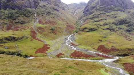 Vista-Aérea-Del-Espectacular-Paisaje-Del-Valle-De-Glencoe-En-Escocia-Con-Un-Río-Que-Fluye-Montaña-Abajo