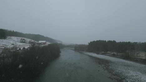 Snowfall-gently-descends-over-a-tranquil-Norwegian-river-surrounded-by-bare-trees,-evoking-a-serene-winter-atmosphere,-wide-shot