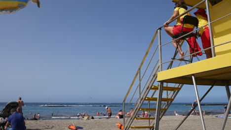 Los-Socorristas-Vigilan-Desde-Una-Torre-Amarilla-En-Una-Vibrante-Playa-Salpicada-De-Bañistas,-La-Extensión-Azul-Del-Mar-Se-Despliega-Bajo-Un-Cielo-Despejado.