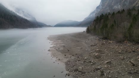 Drone-flies-along-grey-frozen-shoreline-of-reservoir-lake-with-misty-clouds-surrounding-mountains-in-distance