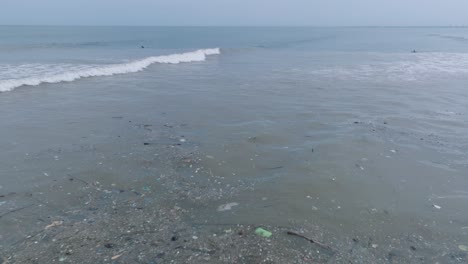 Low-altitude-pan-down-drone-shot-of-polluted-ocean-filled-with-floating-trash-and-debris-and-muddy-sewage-river-runoff-with-surfers-in-the-background-in-Bali-Indonesia
