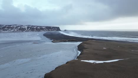 Schwarzer-Sandstrand-Von-Reynisfjara-In-Island-Mit-Reynisdrangar-Klippen-In-Der-Ferne,-Luftaufnahme-Links