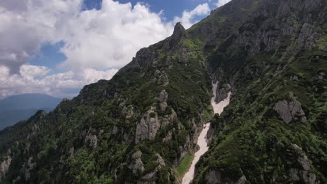 A-muddy-river-flowing-through-bucegi-mountains,-green-peaks-under-a-blue-sky,-summer-day,-aerial-view