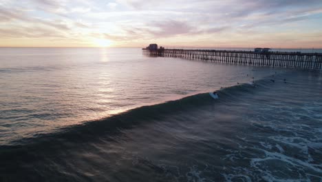 Aerial-drone-view-of-surfers-riding-gentle-waves-near-a-pier-during-a-beautiful-sunset,-capturing-the-essence-of-tranquility-and-leisure