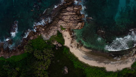 A-top-down-drone-shot-reveals-the-rocky-coastline-of-a-beautiful-island-lined-with-boulders-and-coral-reefs-in-Seychelles,-Africa