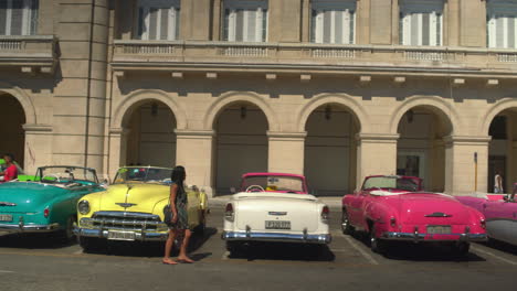 Amidst-Central-Havana's-bustling-Prado,-a-tourist-navigates-past-a-procession-of-vibrant,-classic-1950s-American-cars-and-colorful-buildings-along-the-iconic-Paseo-de-Marti