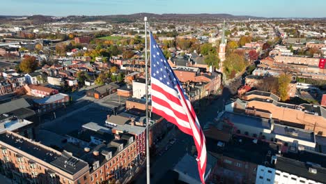 Vista-Aérea-De-York,-Pensilvania,-Con-Una-Bandera-Estadounidense-En-Primer-Plano-Y-La-Arquitectura-Histórica-De-La-Ciudad-Debajo.