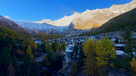 Early-morning-Saas-Fee-Zermatt-Saastal-first-light-sunny-Swiss-alps-mountain-peaks-shaded-alpine-valley-chalet-ski-resort-town-Switzerland-downtown-river-crossway-bridge-slow-pan-to-the-left