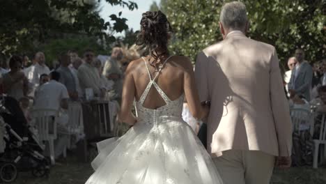 Desde-Atrás,-Un-Padre-Lleva-A-Su-Hija-Por-El-Pasillo-Con-Familiares-Y-Amigos-A-Ambos-Lados,-Ceremonia-De-Boda-Al-Aire-Libre-Sobre-El-Césped-Con-árboles-Alrededor