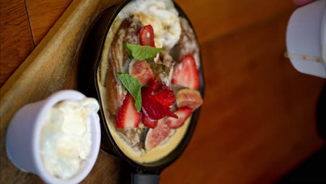 Vertical-slow-motion-of-a-person-pouring-a-fruit-sauce-onto-a-cinnamon-roll-topped-with-sliced-figs-and-strawberries-in-an-iron-skillet