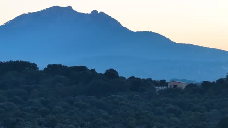 Vuelo-De-Madrugada-Visualizando-En-166mm-Una-Finca-Con-Una-Casa-Rural-De-Piedra-En-Construcción-Con-El-Fondo-De-Una-Montaña-Creando-Un-Efecto-De-Paralaje-Con-Un-Cielo-Blanquecino-En-Invierno-Avila-España