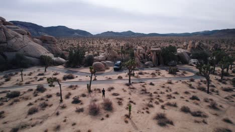Una-Camioneta-Mercedes-Sprinter-Estacionada-En-El-Parque-Nacional-Joshua-Tree-Mientras-Un-Dron-Vuela-Hacia-Atrás-Desde-Allí,-En-El-Video-Se-Ven-Escarpadas-Formaciones-Rocosas-Y-árboles-De-Joshua