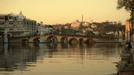 Featuring-the-Chandpole-bridge-in-Lake-Pichola-in-Udaipur-India,-with-a-mesmerizing-backdrop-of-beautiful-white-architecture-in-the-background-just-after-sunset