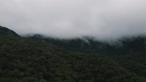 -Ominous-weather-wit-low-clouds-collecting-on-the-mountainside-creating-a-mysterious-atmosphere-over-a-dense-forest,-aerial-drone-footage