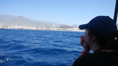 Contemplative-pensive-woman-on-a-boat,-viewing-a-distant-cityscape-across-the-sea,-with-mountains-under-a-clear-sky