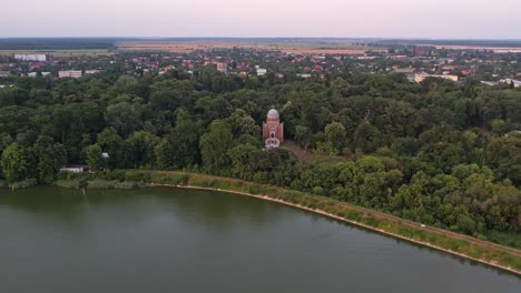 A-historic-church-surrounded-by-greenery-near-a-lake-at-dawn,-aerial-view