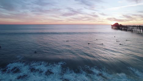 Eine-Ruhige-Meereslandschaft-Bei-Sonnenuntergang-Am-Kalifornischen-Meer-Mit-Einem-Sich-Abzeichnenden-Pier-Am-Meer-Und-Surfern,-Die-Wellen-Fangen,-Aufgenommen-Mit-Einer-Drohnen-Luftaufnahme