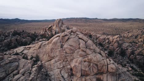 Drones-Volando-En-El-Parque-Nacional-Joshua-Tree-Sobre-Formaciones-Rocosas-Escarpadas-Mientras-Un-Hombre-Solitario-Se-Encuentra-En-La-Cima-De-La-Roca