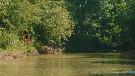 Man-in-red-casting-a-fishing-net-into-a-tranquil-river-in-Arauca,-Colombia,-surrounded-by-lush-greenery,-slow-motion