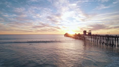 Un-Tranquilo-Paisaje-Marino-Al-Atardecer-En-Oceanside,-California,-Con-Siluetas-De-Muelle-Oceánico-Y-Surfistas-Capturando-Olas-Capturadas-Con-Una-Toma-Aérea-De-Drones