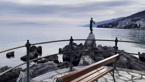 Estatua-De-Una-Mujer-En-El-Banco-De-La-Orilla-Del-Mar-Ciudad-Turística-Isla-En-La-Distancia-Lovran,-Opatija,-Croacia