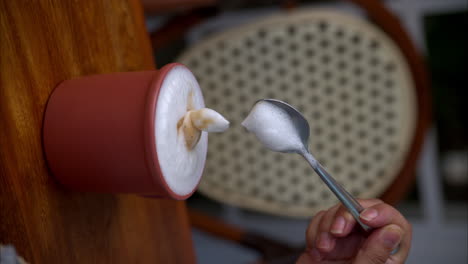 Vertical-slow-motion-of-a-female-hand-playing-with-the-foam-of-a-cappuccino-using-a-metal-spoon