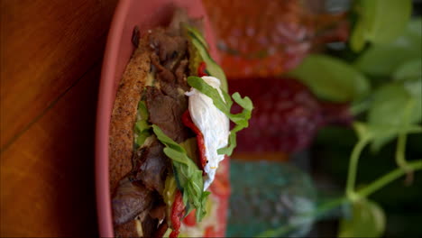 Vertical-Slow-motion-pullback-shot-of-a-beef-deli-sandwich-topped-with-arugula-mozzarella-cheese-pickles-cucumber-and-tomato-served-with-salad