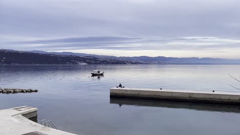 Fisherman-casting-a-line-on-peer-Boat-parked-on-Sea-shore-with-town-in-distance-wide,-Opatija,-Croatia