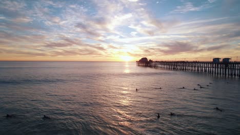 Eine-Ruhige-Meereslandschaft-Bei-Sonnenuntergang-Am-Kalifornischen-Meer-Mit-Einem-Sich-Abzeichnenden-Pier-Am-Meer-Und-Surfern,-Die-Wellen-Fangen,-Aufgenommen-Mit-Einer-Drohnen-Luftaufnahme