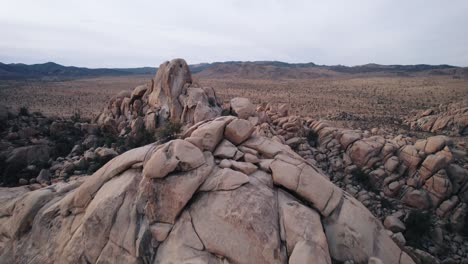 Drone-flying-in-Joshua-Tree-National-Park-over-rugged-rock-formations-while-a-lonely-man-standing-on-top-of-the-rock