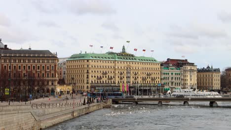 Grand-Hotel-overlooks-stream-as-buses-cross-bridge,-flags-fly-on-roof