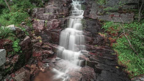 Ein-Zeitraffervideo-Eines-Kleinen-Wasserfalls,-Der-über-Die-Dunklen-Felsen-Im-Sommerwald-Stürzt