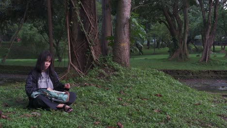 Static-shot-of-teenager-girl-sitting-down-and-drawing-on-sketchbook-in-forest-park,-gets-up-and-walks-away