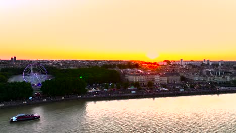 Sonnenuntergang-Hinter-überfüllten-Weinmesse-Mit-Beleuchtetem-Riesenrad-Mit-Fluss-Garonne-Und-Boot,-Luftaufnahme-Von-Links