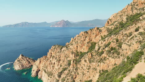 Rugged-Corsica-coastline-with-clear-blue-sea,-golden-sunlight,-and-mountainous-terrain,-aerial-shot
