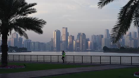 Ein-Bewohner-Genießt-Einen-Morgendlichen-Spaziergang-An-Der-Corniche-Von-Sharjah-Mit-Blick-Auf-Die-Skyline-Von-Sharjah-Im-Hintergrund.