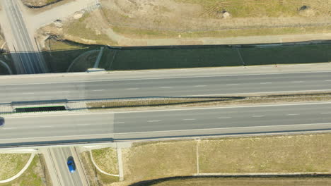 Aerial-view-of-a-dual-carriageway-with-a-single-white-car-traveling-on-the-right-lane,-amidst-expansive-fields-under-a-clear-sky