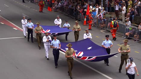 Soldaten-Der-Drei-Zweige-Der-Australischen-Streitkräfte,-Der-Royal-Australian-Navy,-Der-Australian-Army-Und-Der-Royal-Australian-Air-Force,-Halten-Die-Flaggen-Und-Marschieren-Bei-Der-Anzac-Day-Parade-Die-Straße-Entlang