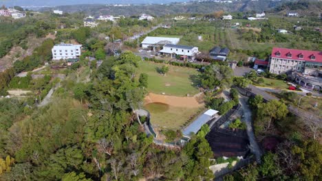 A-lush,-green-rural-landscape-with-scattered-buildings-and-a-small-pond,-under-clear-skies,-aerial-view